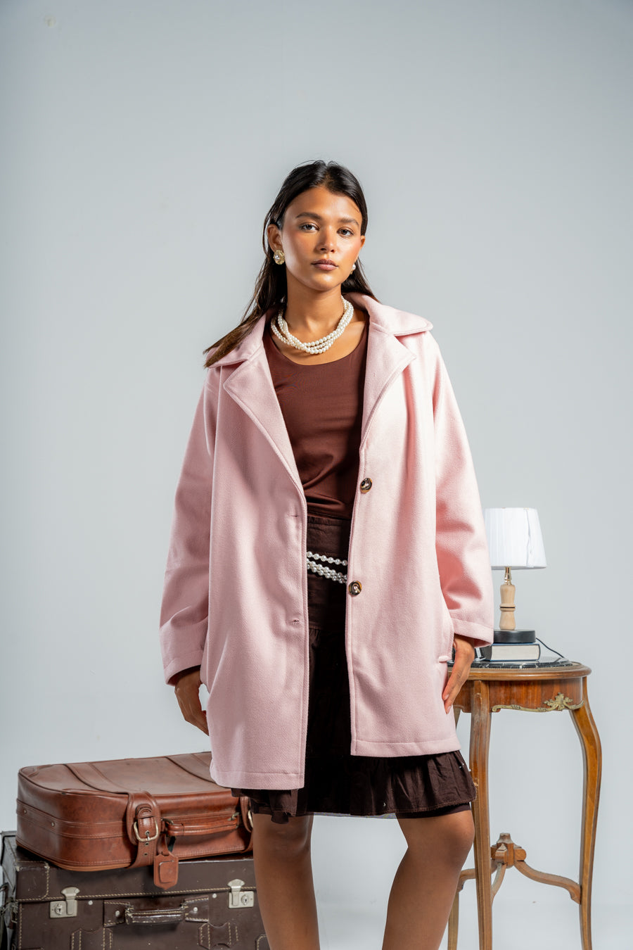 Woman wearing a pink coat over a brown dress, standing next to vintage suitcases and a small table with a lamp.shop_this_look_86DbjN