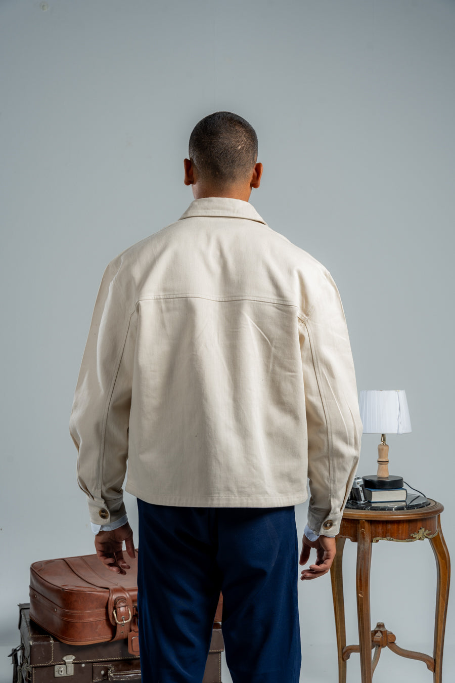 Man wearing a beige jacket and blue pants standing next to vintage suitcases and a table with a lamp.
