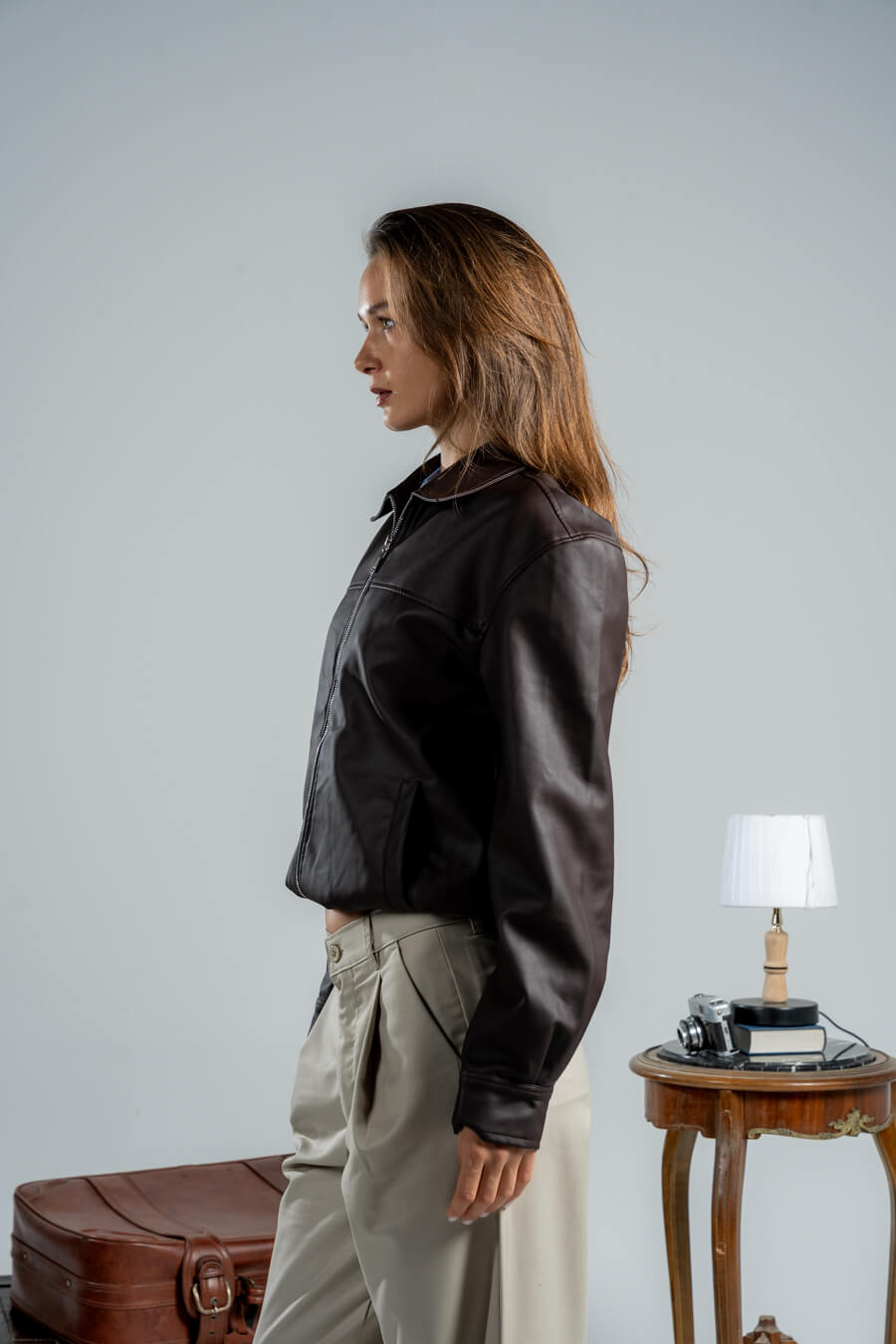 Woman wearing a dark jacket and light pants standing next to a table with a lamp and suitcase.