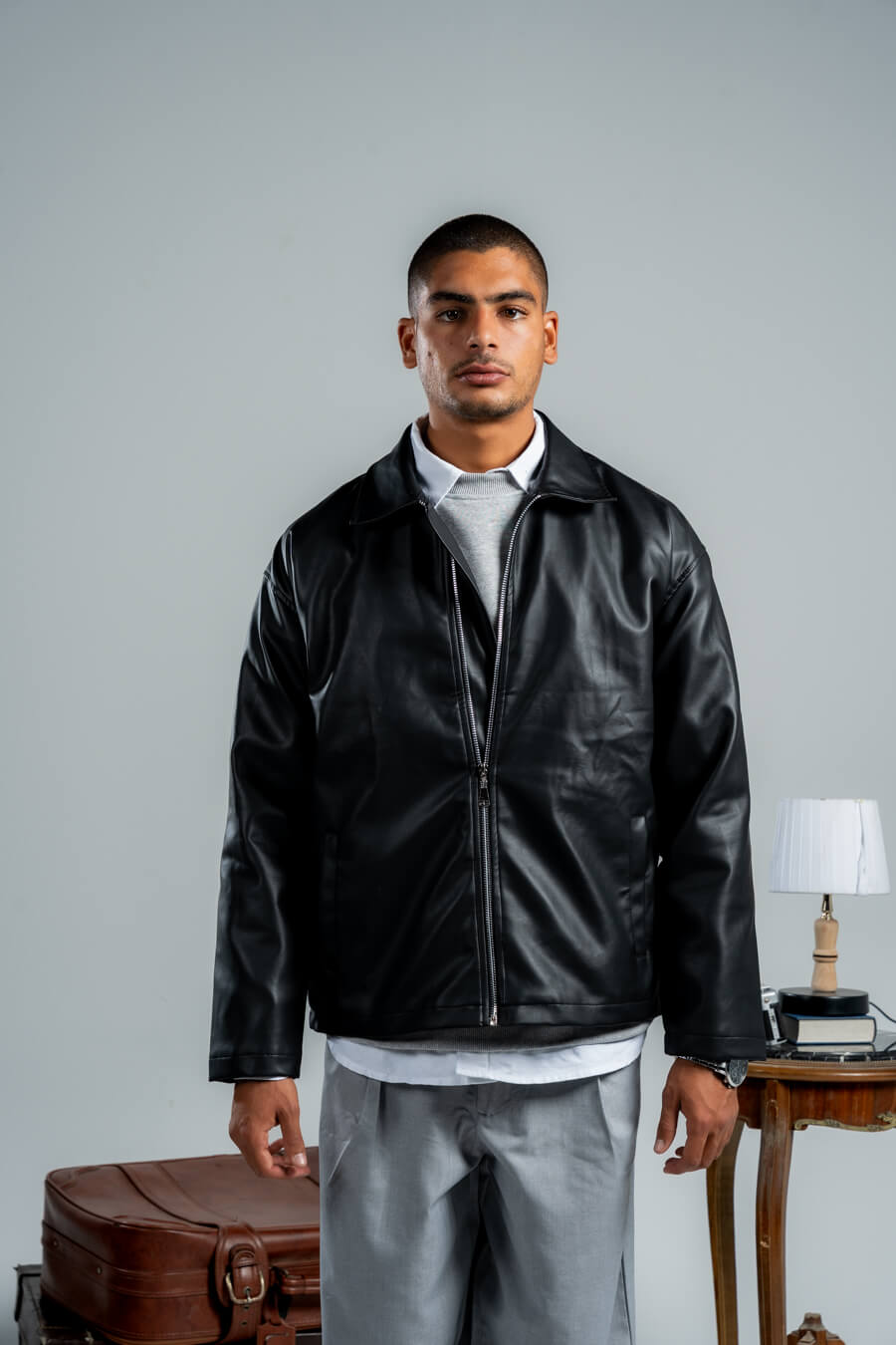 Man wearing a black leather jacket standing in a room with a table and lamp.