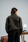 Person wearing a green jacket and black pants standing in front of a plain wall with a suitcase and table in the background.
