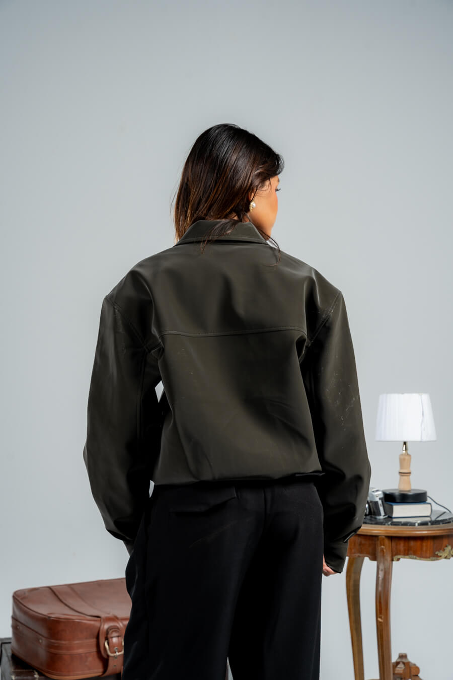 Person wearing a green jacket and black pants standing in front of a plain wall with a suitcase and table in the background.