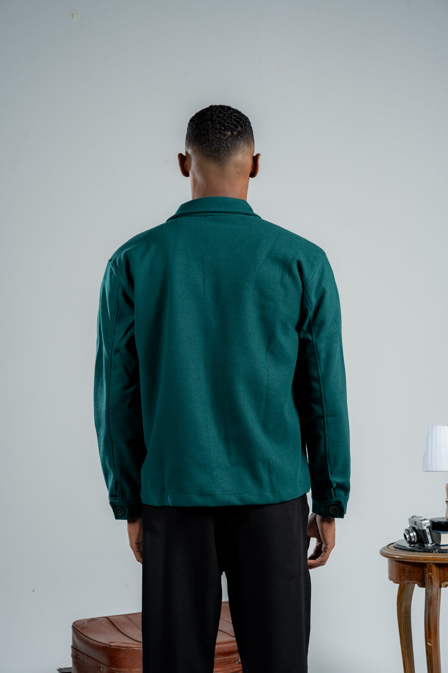 Person wearing a green shirt and black pants standing against a plain wall with a small table and lamp in the background.