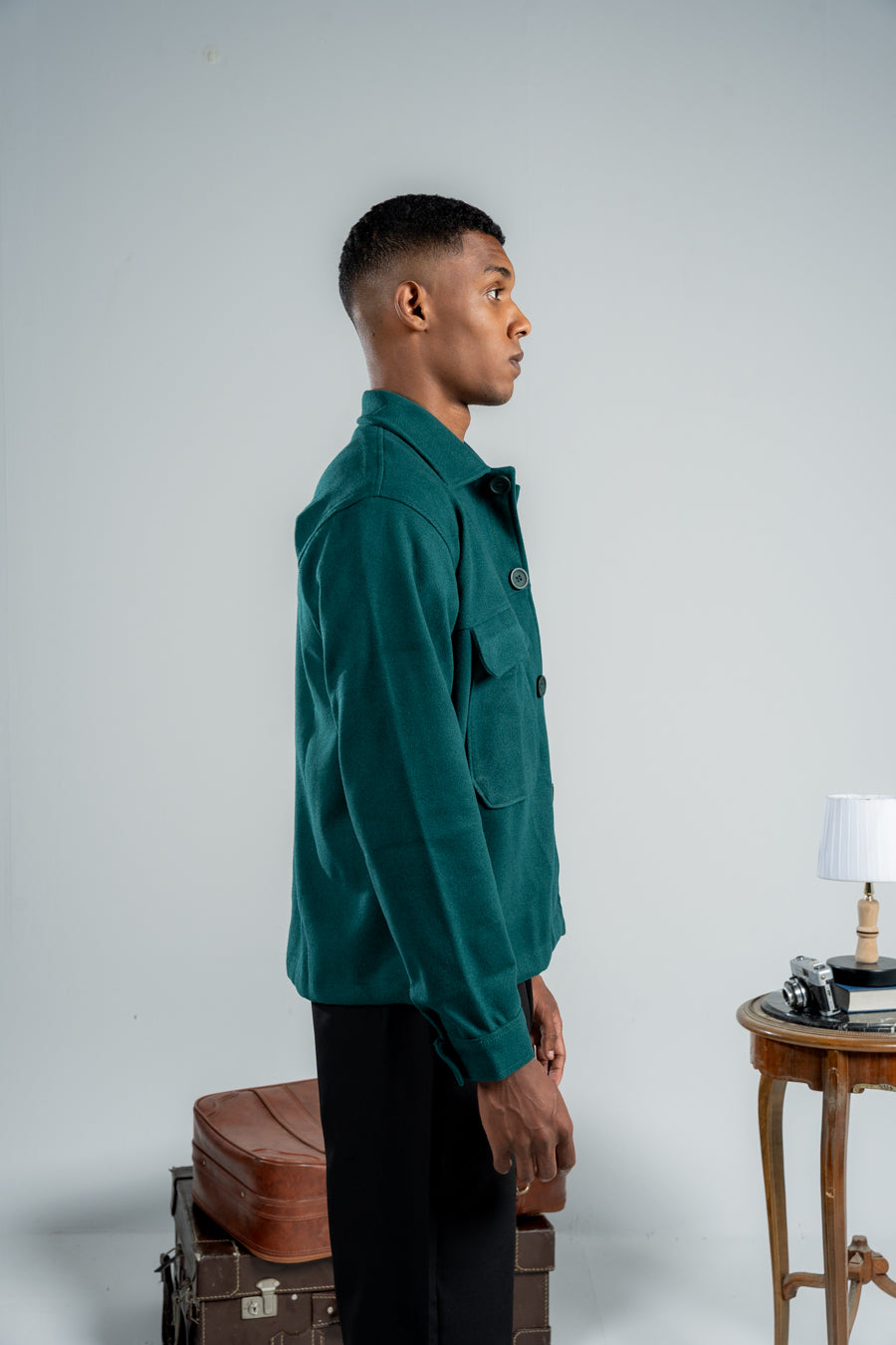 Man wearing a green jacket standing next to vintage suitcases and a small table with a lamp against a plain background
