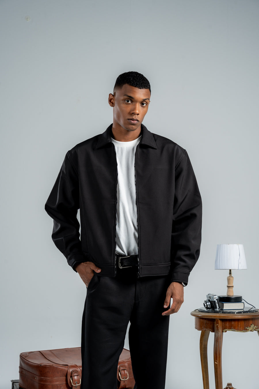 Man wearing a black jacket and pants standing in a room with a table and lamp.