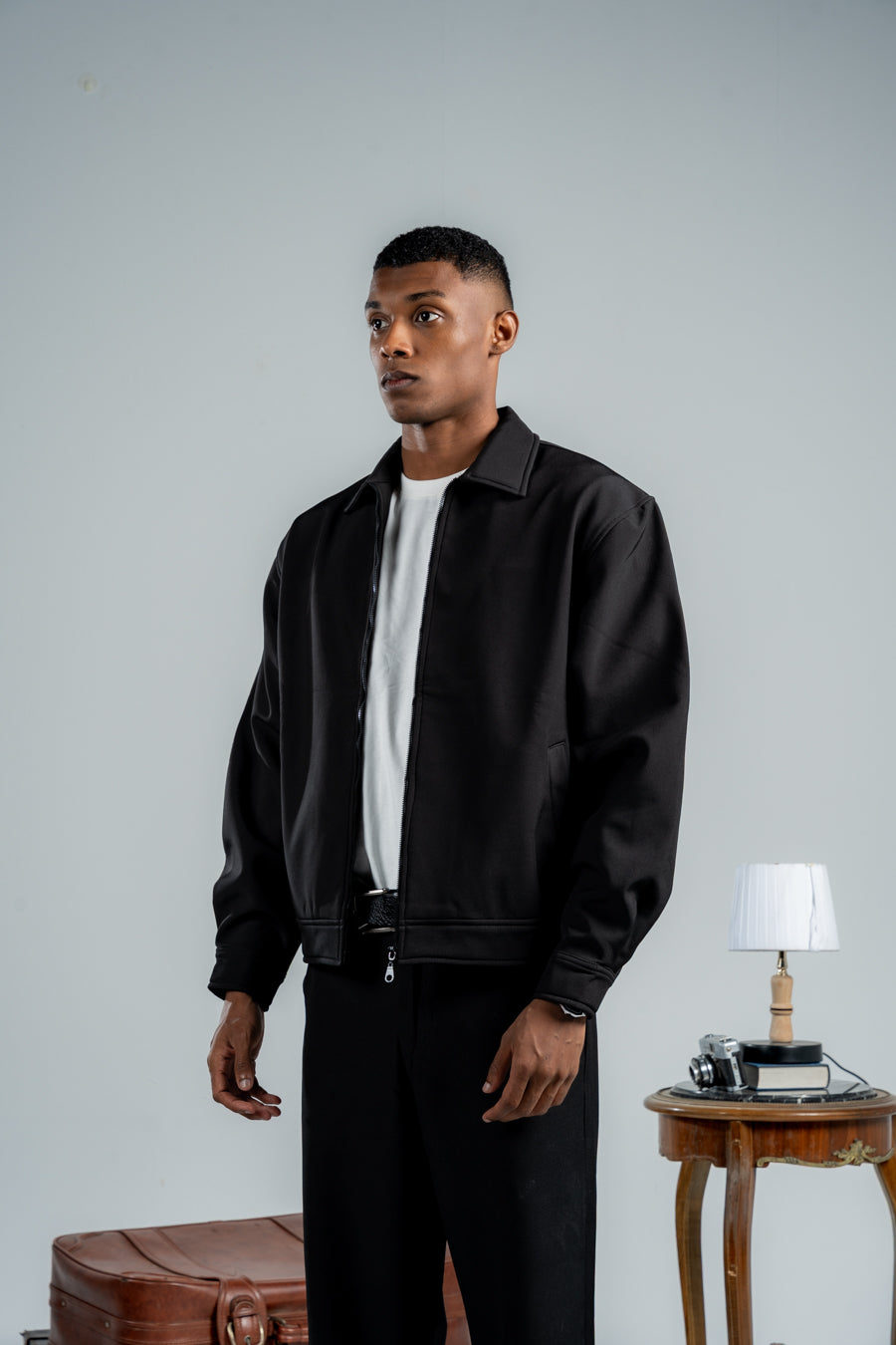 Man wearing a black jacket and pants standing in a room with a table and lamp.