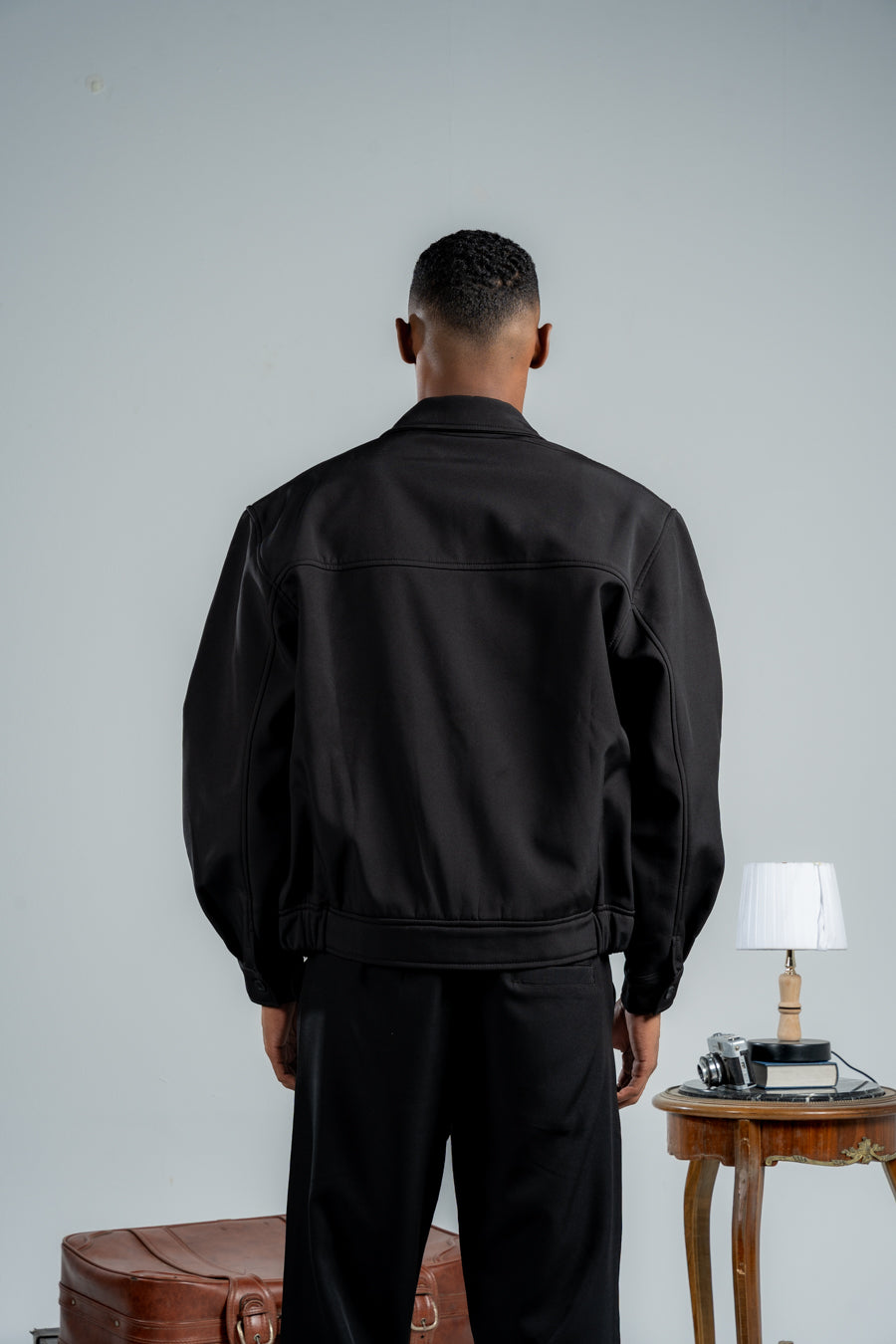 Man wearing a black jacket standing in front of a plain wall with a small table and lamp in the background.