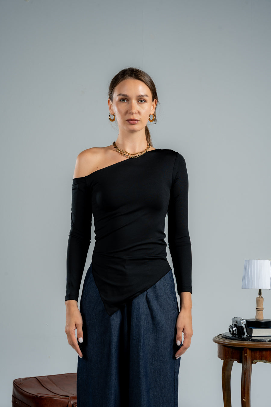 Woman wearing a black one-shoulder top and blue pants against a gray background