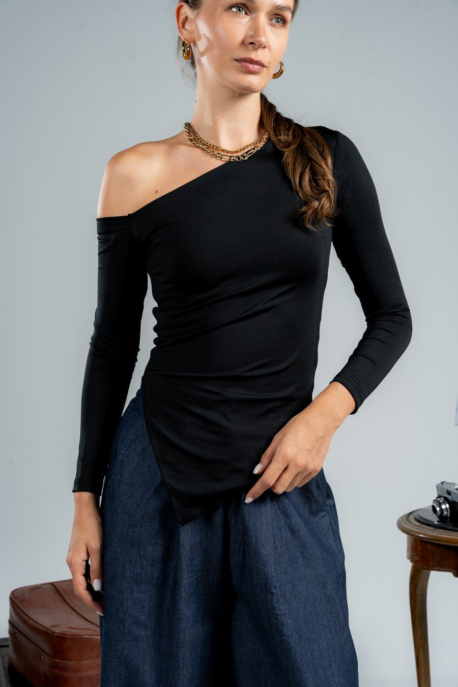 Woman wearing a black one-shoulder top and blue pants against a gray background