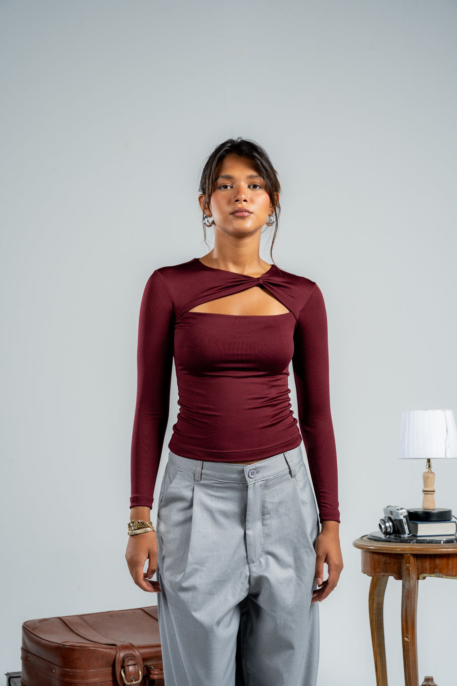 Woman wearing a burgundy long-sleeve top with cut-out design and light gray pants, standing against a plain background.