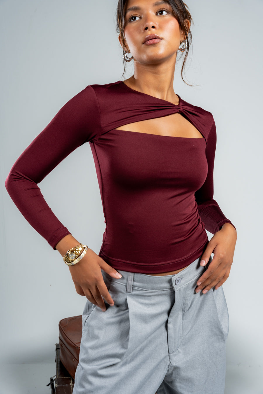 Woman wearing a burgundy long-sleeve top with cut-out design and light gray pants, standing against a plain background.