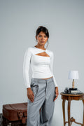 Woman wearing a white long-sleeve top with cut-out design and gray pants, standing next to a suitcase and small table.
