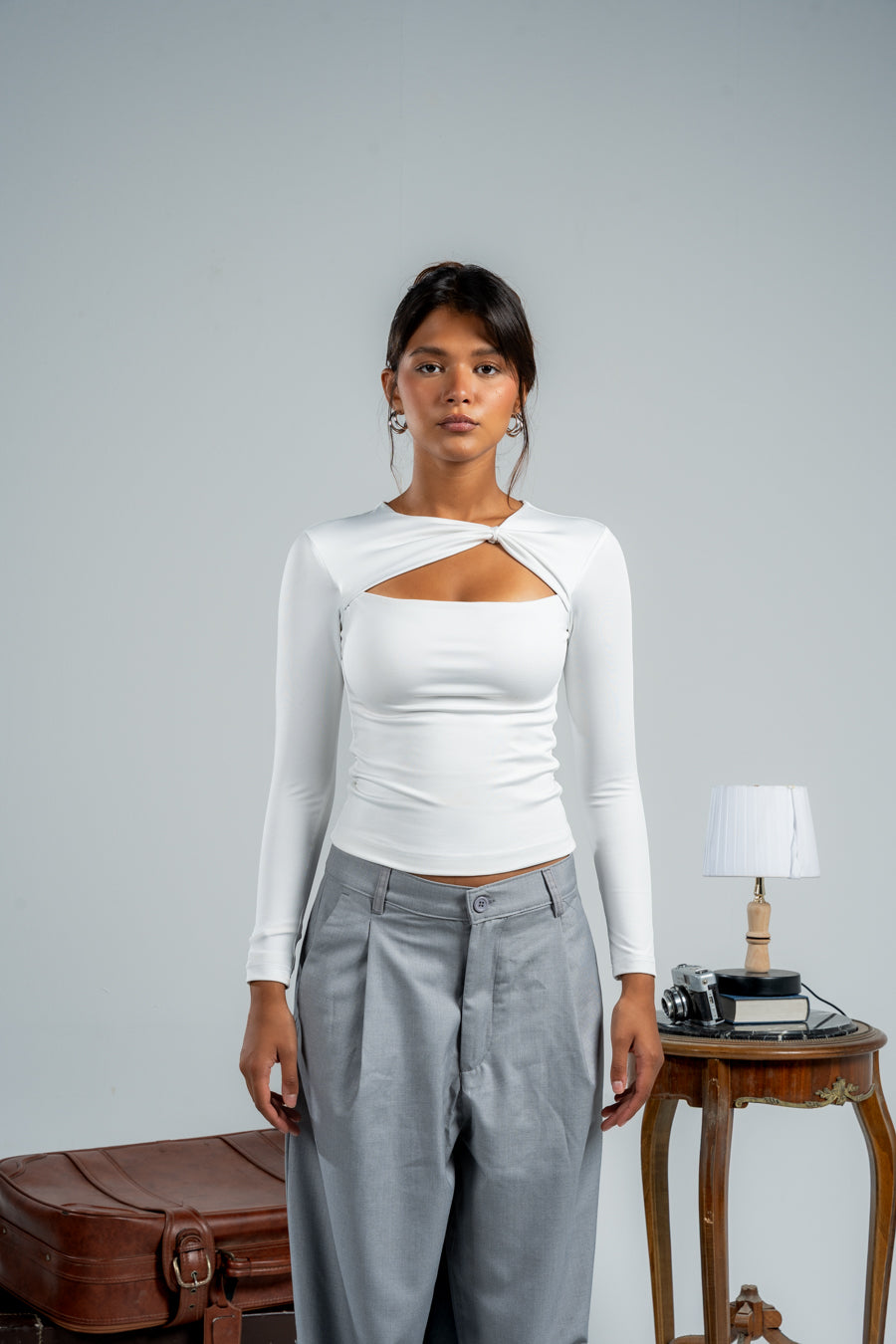 Woman wearing a white long-sleeve top with a cutout design and gray pants against a plain background.