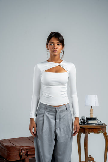 Woman wearing a white long-sleeve top with a cutout design and gray pants against a plain background.