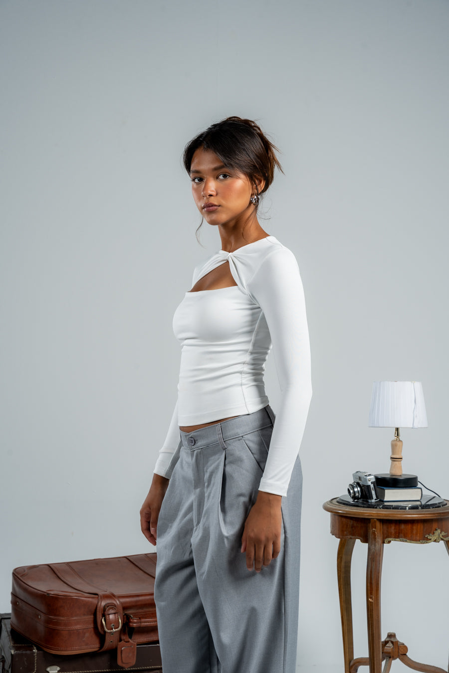 Woman in a white top and gray pants standing next to a suitcase against a plain background