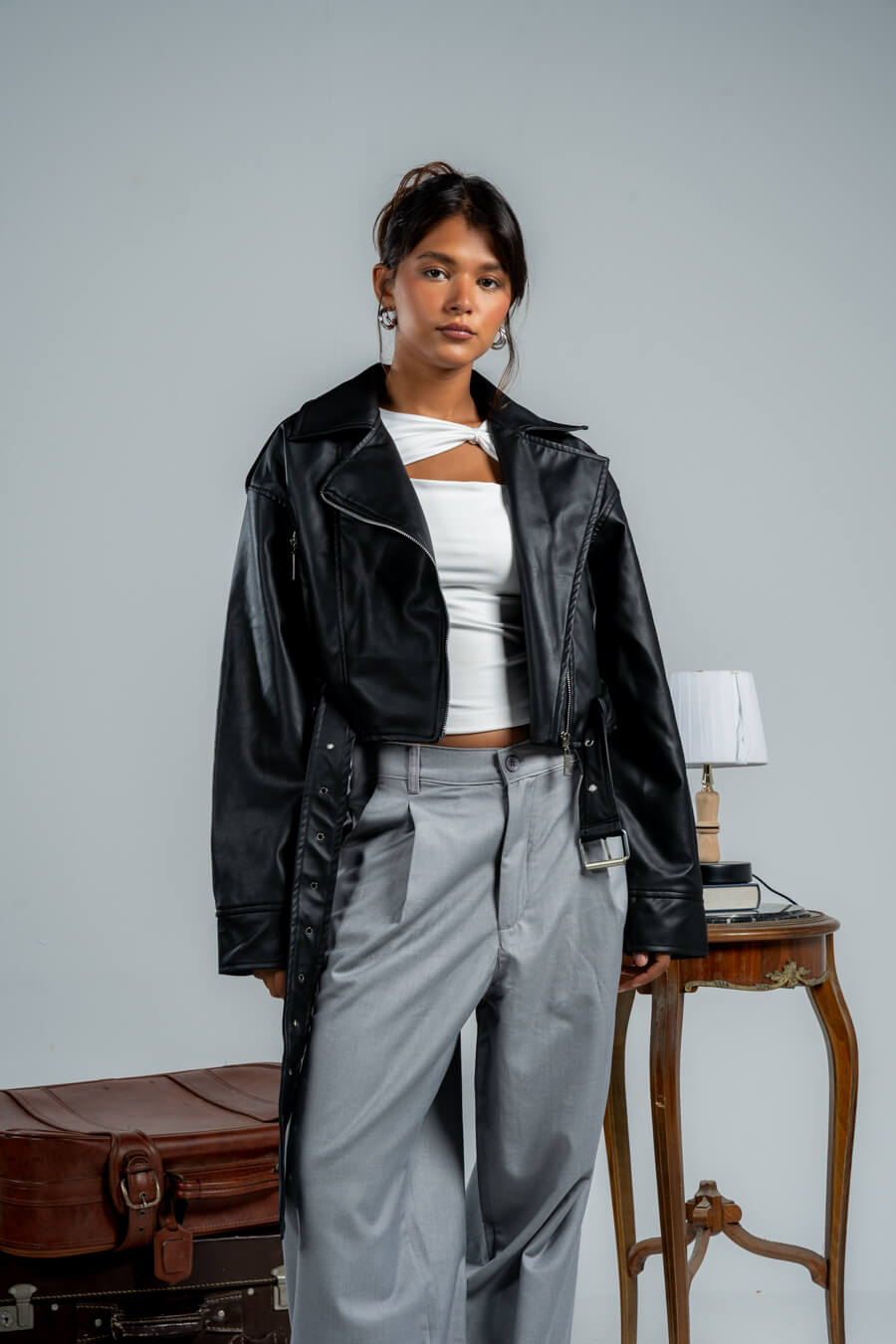 Person wearing a black leather jacket over a white top and gray pants, standing in a room with a suitcase and table.