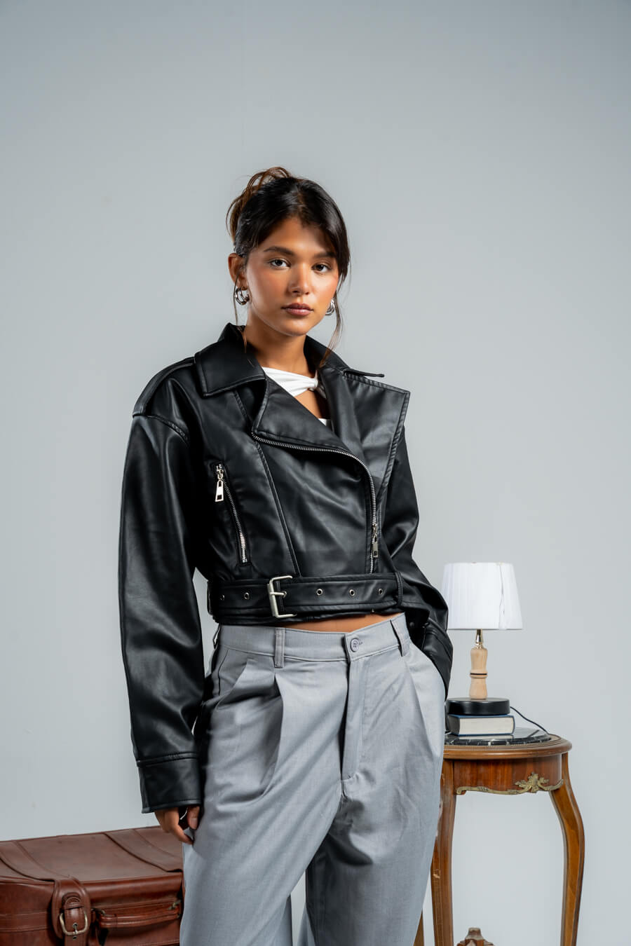 Person wearing a black leather jacket over a white top and gray pants, standing in a room with a suitcase and table.