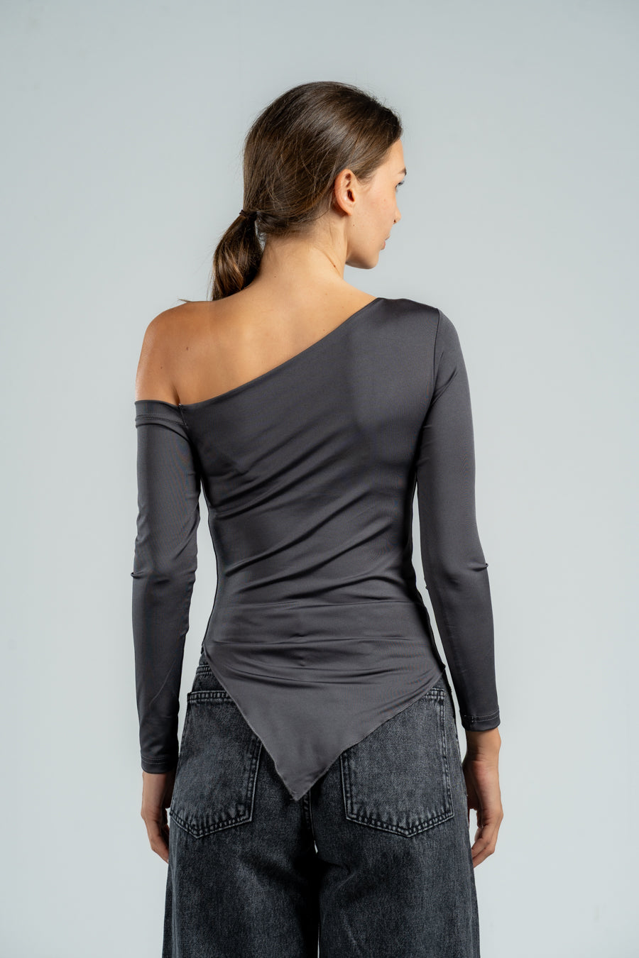 Woman wearing a gray one-shoulder top and dark jeans against a light gray background