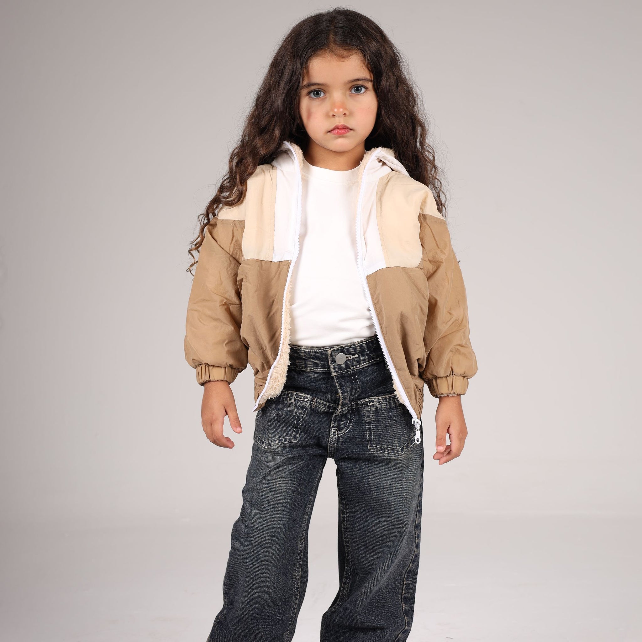 Child wearing a beige jacket, white shirt, and dark jeans on a plain background