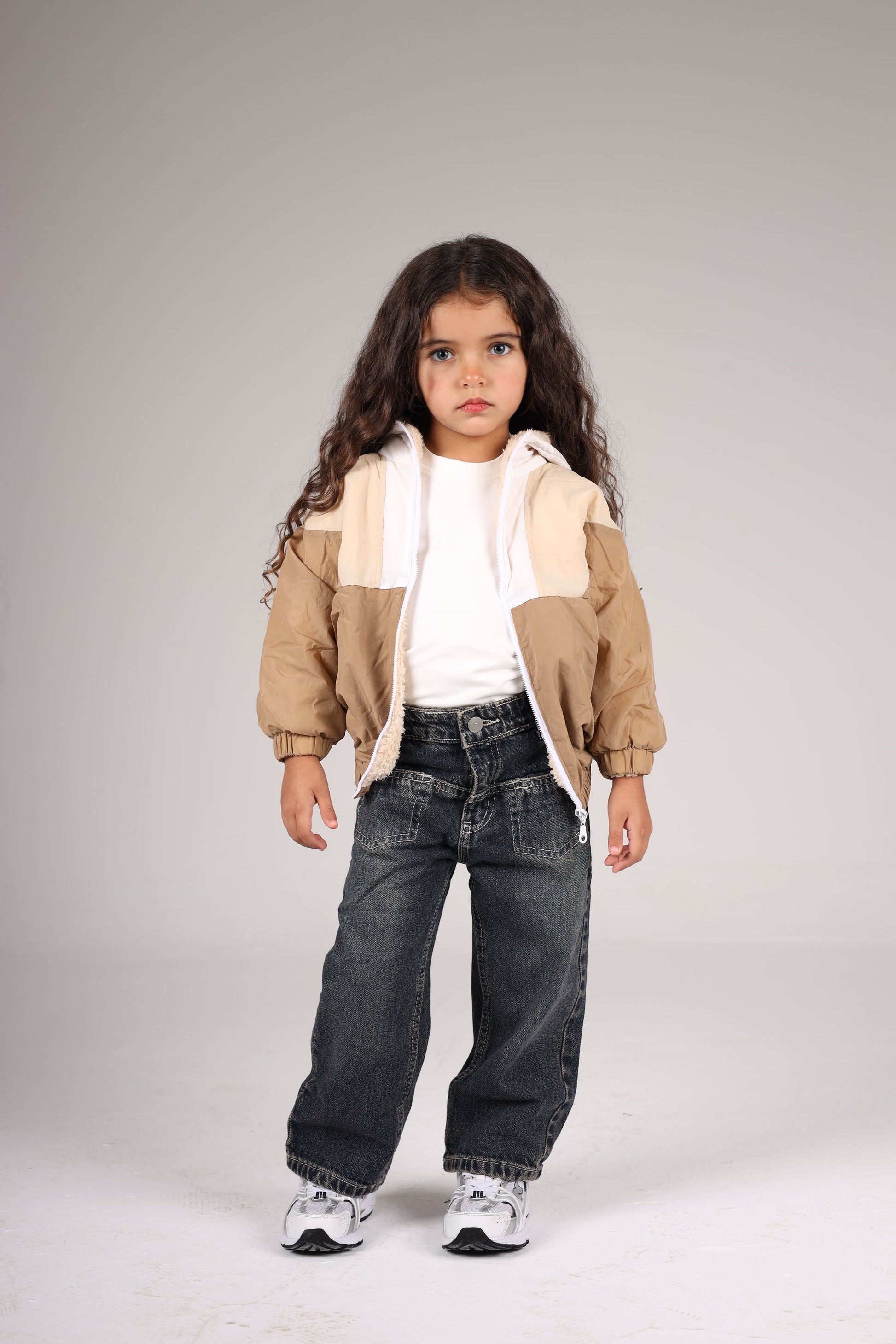 Child wearing a beige jacket, white shirt, and dark jeans on a plain background