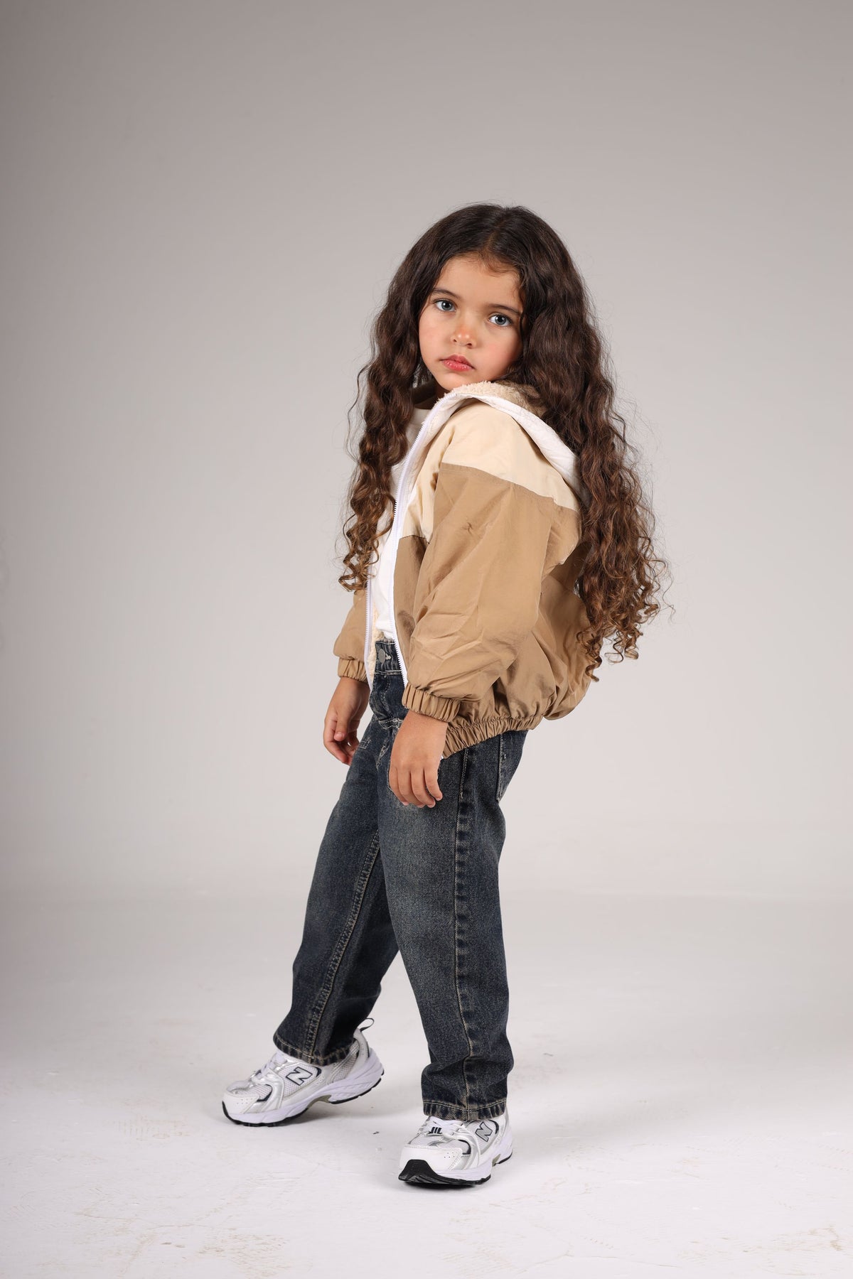 Young girl wearing a beige jacket, white shirt, blue jeans, and white sneakers on a plain background