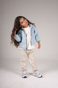 Child wearing a denim jacket, white shirt, beige pants, and sneakers on a plain background