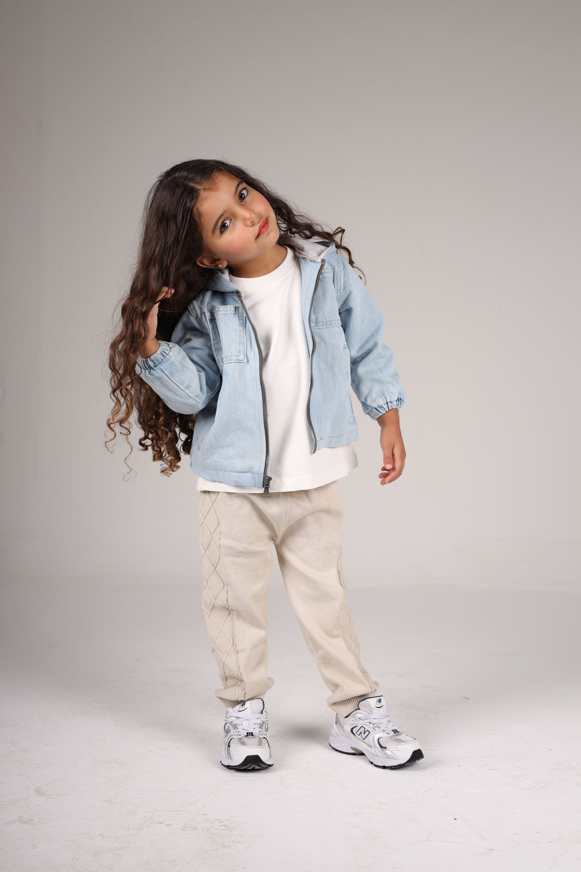 Child wearing a denim jacket, white shirt, beige pants, and sneakers on a plain background