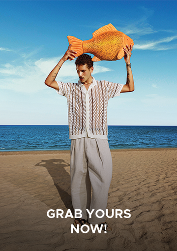A man standing on a sandy beach with the ocean and blue sky behind him, holding a large orange fish-shaped object above his head. He is wearing a white short-sleeve button-up shirt with a brown geometric pattern and light gray pants.