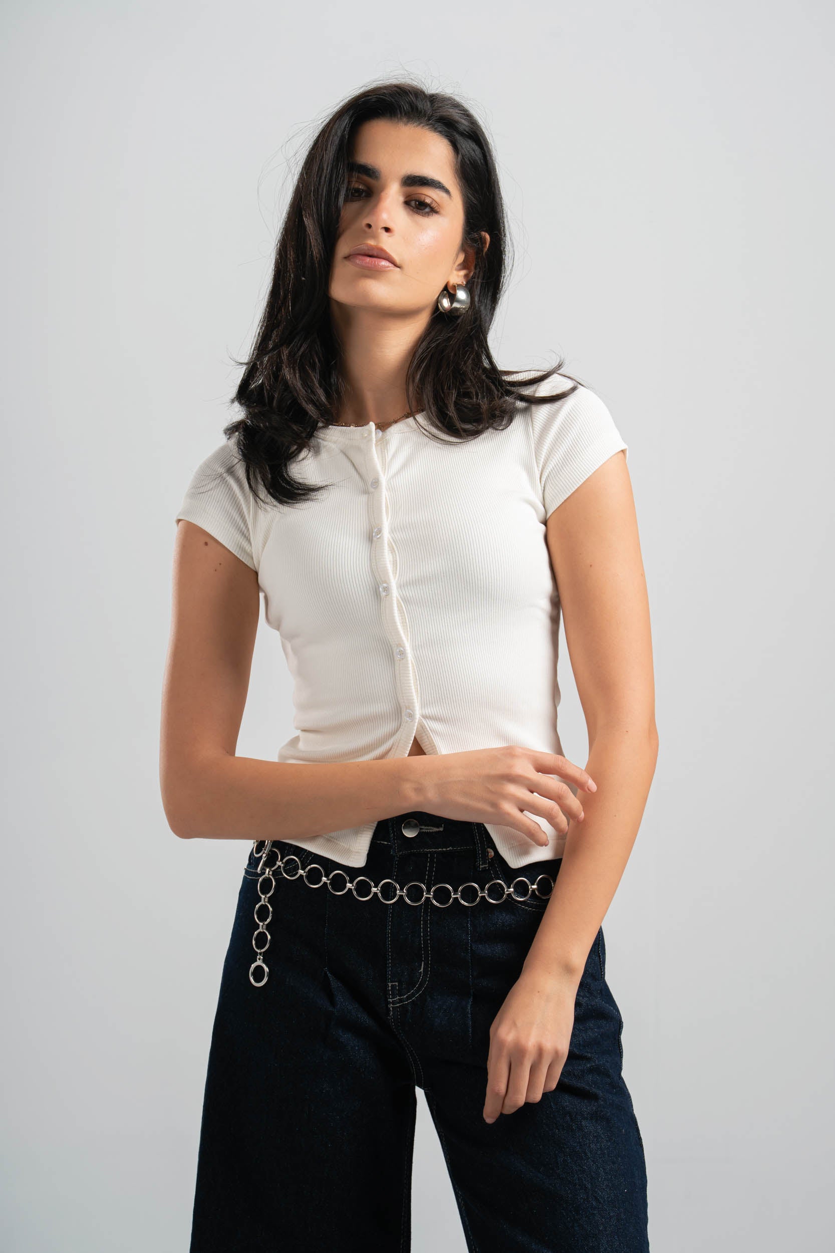 Woman wearing a white blouse and dark jeans with a chain belt against a plain background