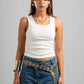 Woman wearing a white tank top and blue jeans with a gold belt against a gray background