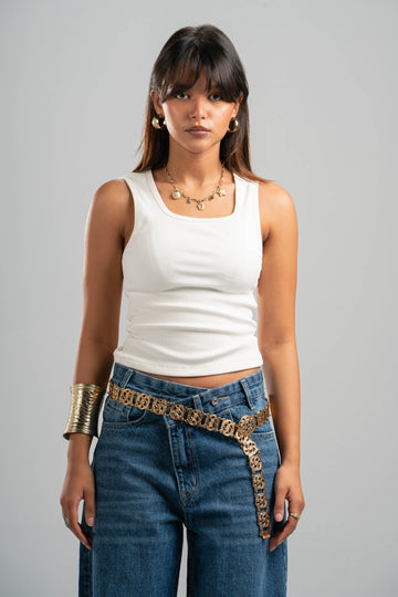 Woman wearing a white tank top and blue jeans with a gold belt against a gray background