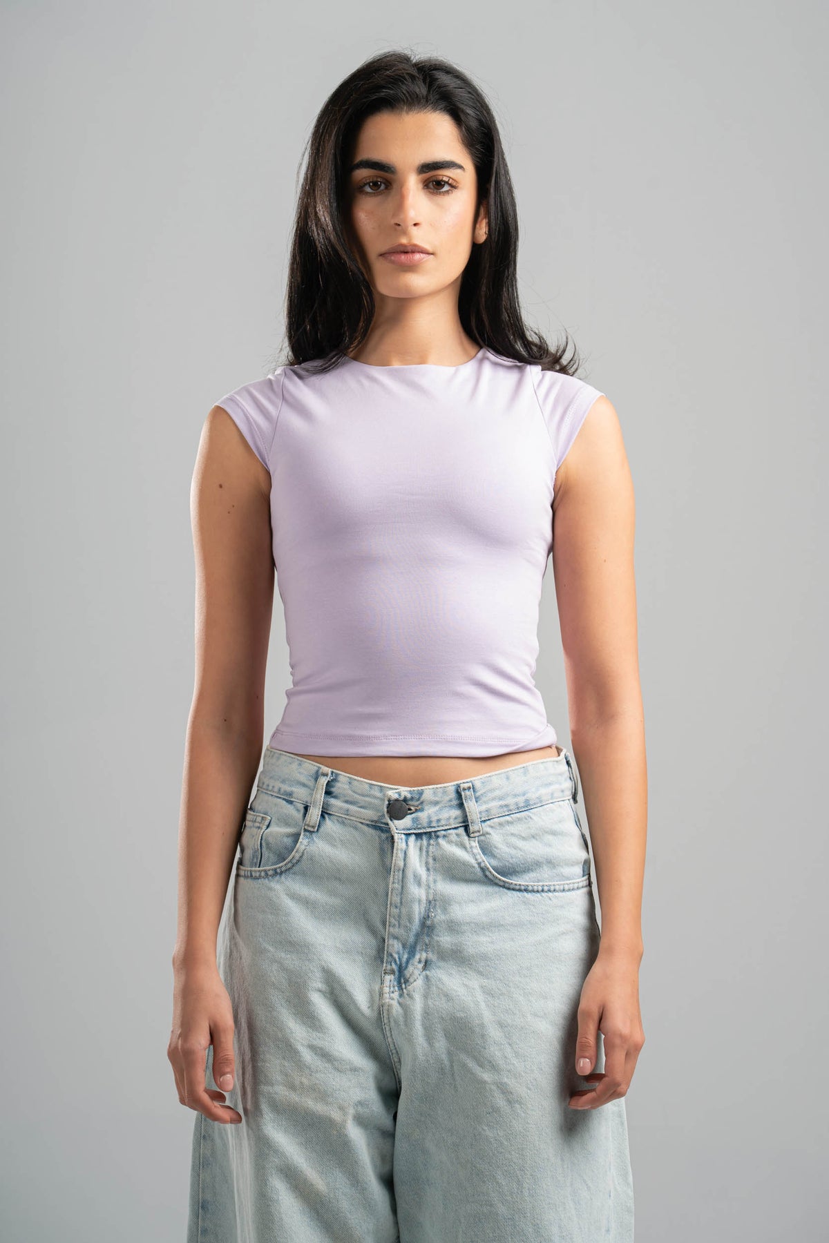 Woman wearing a light purple sleeveless top and light blue jeans against a gray background
