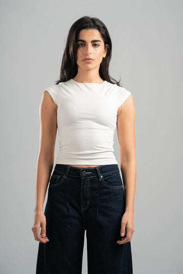 Woman wearing a white sleeveless top and dark jeans against a gray background