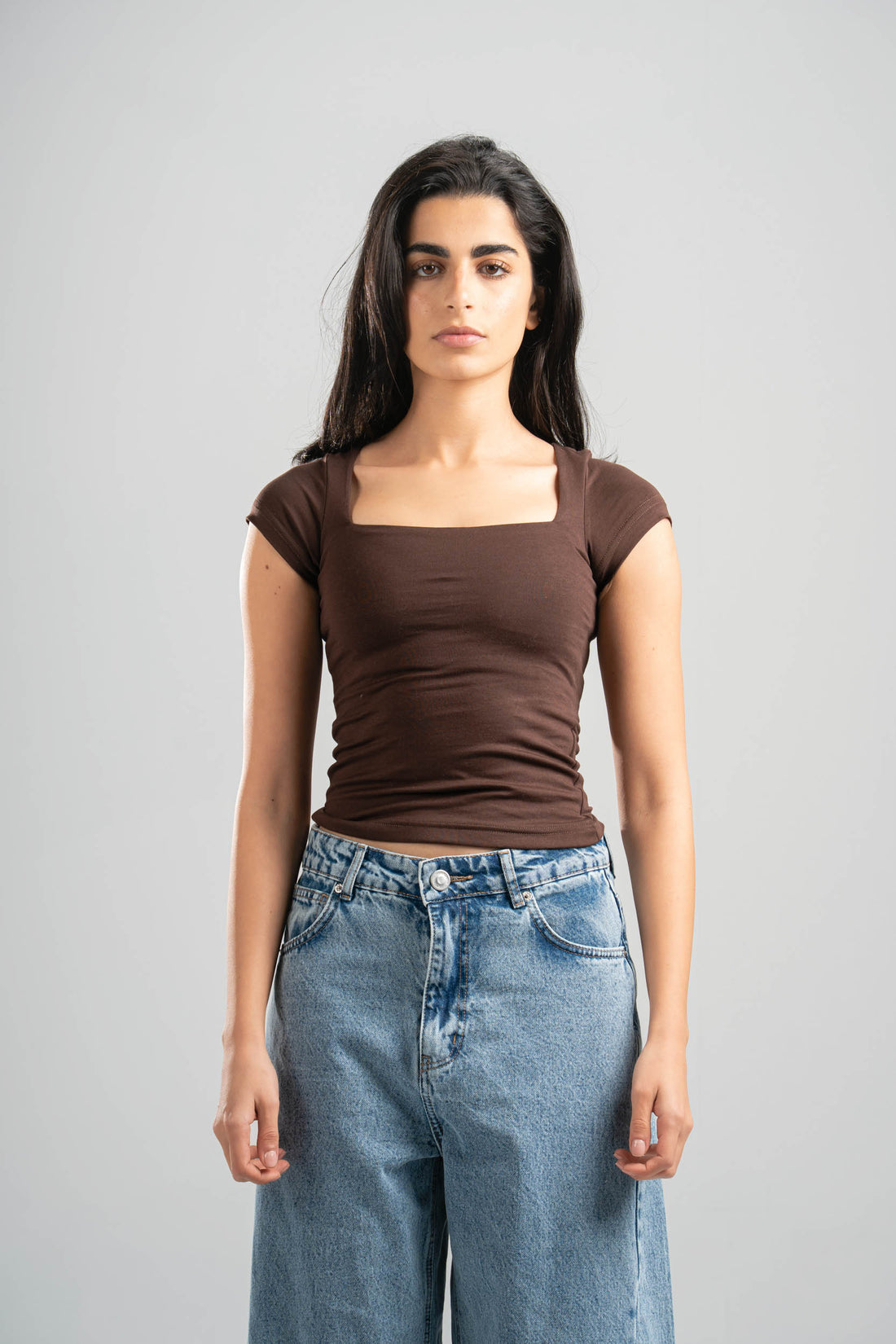 Woman wearing a brown top and blue jeans against a gray background
