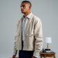 Man in a beige jacket and blue pants standing next to a suitcase with a lamp and table in the background.