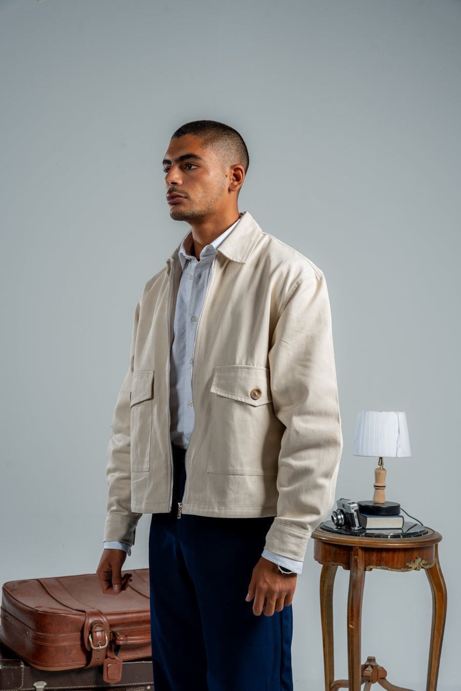 Man in a beige jacket and blue pants standing next to a suitcase with a lamp and table in the background.