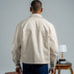 Man wearing a beige jacket and blue pants standing next to vintage suitcases and a table with a lamp.