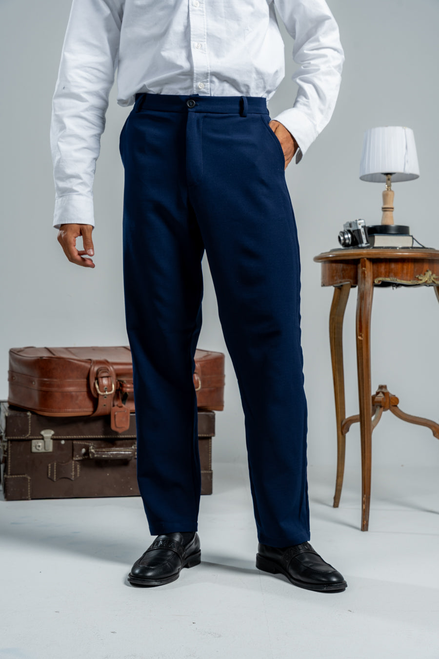 Person wearing a white shirt and navy pants standing in front of vintage suitcases and a small table.