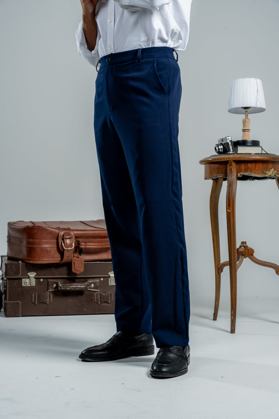 Person wearing navy pants with a white shirt, standing next to vintage suitcases and a small table.