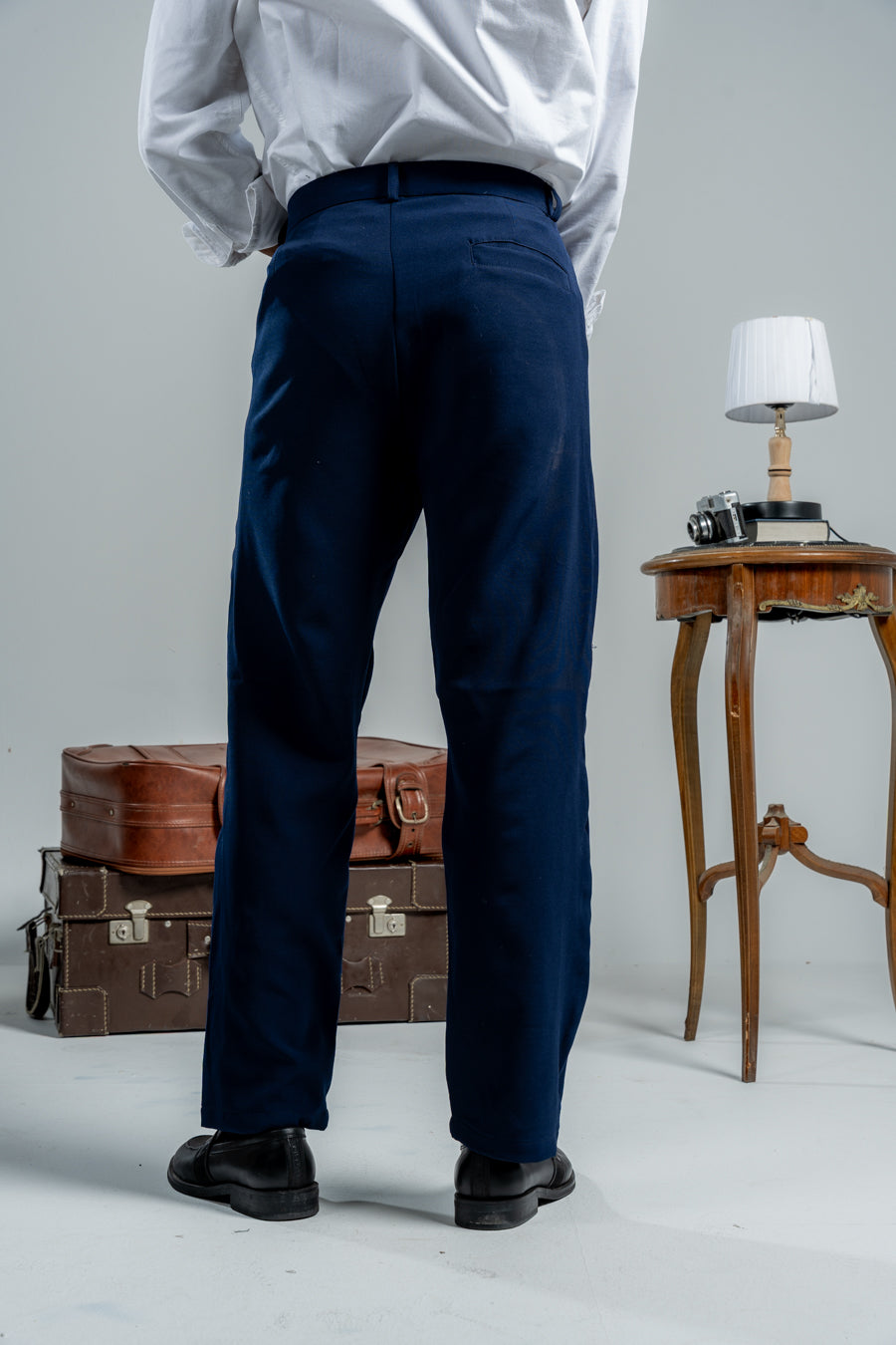 Person wearing navy pants and a white shirt with a gray background, standing next to vintage suitcases and a small table.
