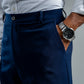 Person wearing a light gray shirt, navy pants, and a watch on a neutral background