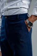 Person wearing a light gray shirt, navy pants, and a watch on a neutral background