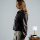 Woman wearing a dark jacket and light pants standing next to a table with a lamp and suitcase.