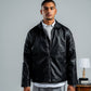 Man wearing a black leather jacket standing in a room with a table and lamp.