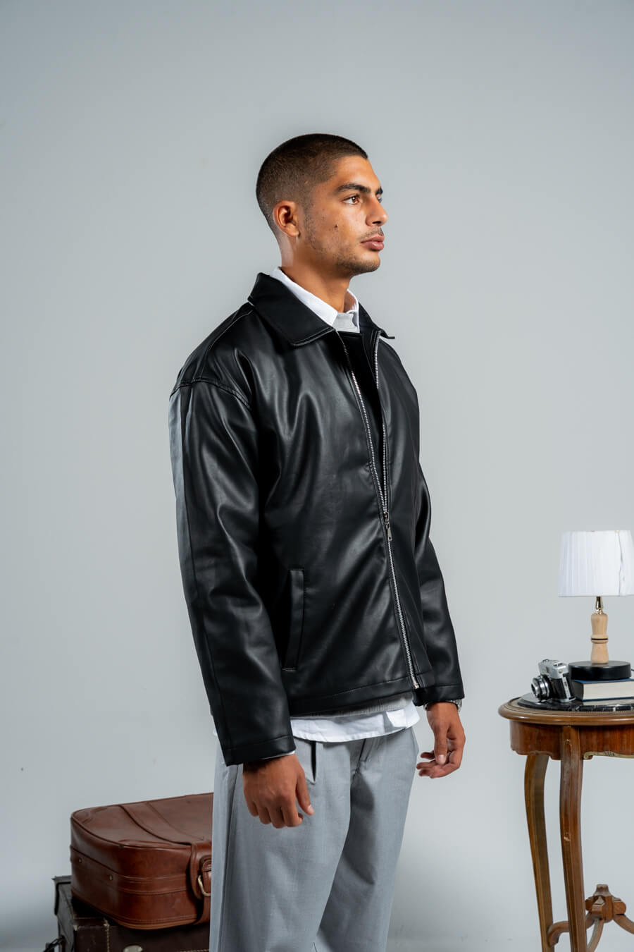 Man wearing a black leather jacket standing next to a suitcase and small table with lamp against a gray background