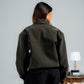 Person wearing a green jacket and black pants standing in front of a plain wall with a suitcase and table in the background.