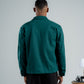 Person wearing a green shirt and black pants standing against a plain wall with a small table and lamp in the background.