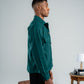 Man wearing a green jacket standing next to vintage suitcases and a small table with a lamp against a plain background