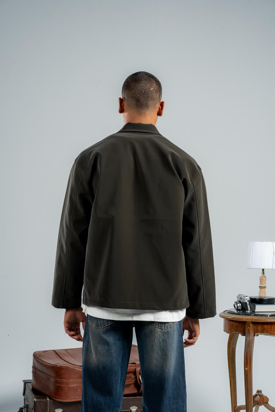 Man wearing a dark green jacket and blue jeans standing in a room with a gray wall and wooden table.