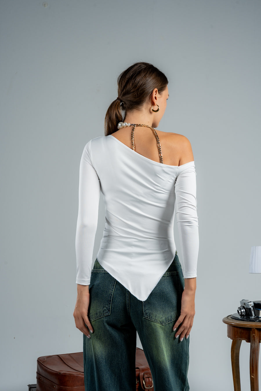 Woman wearing a white one-shoulder top and blue jeans against a gray background